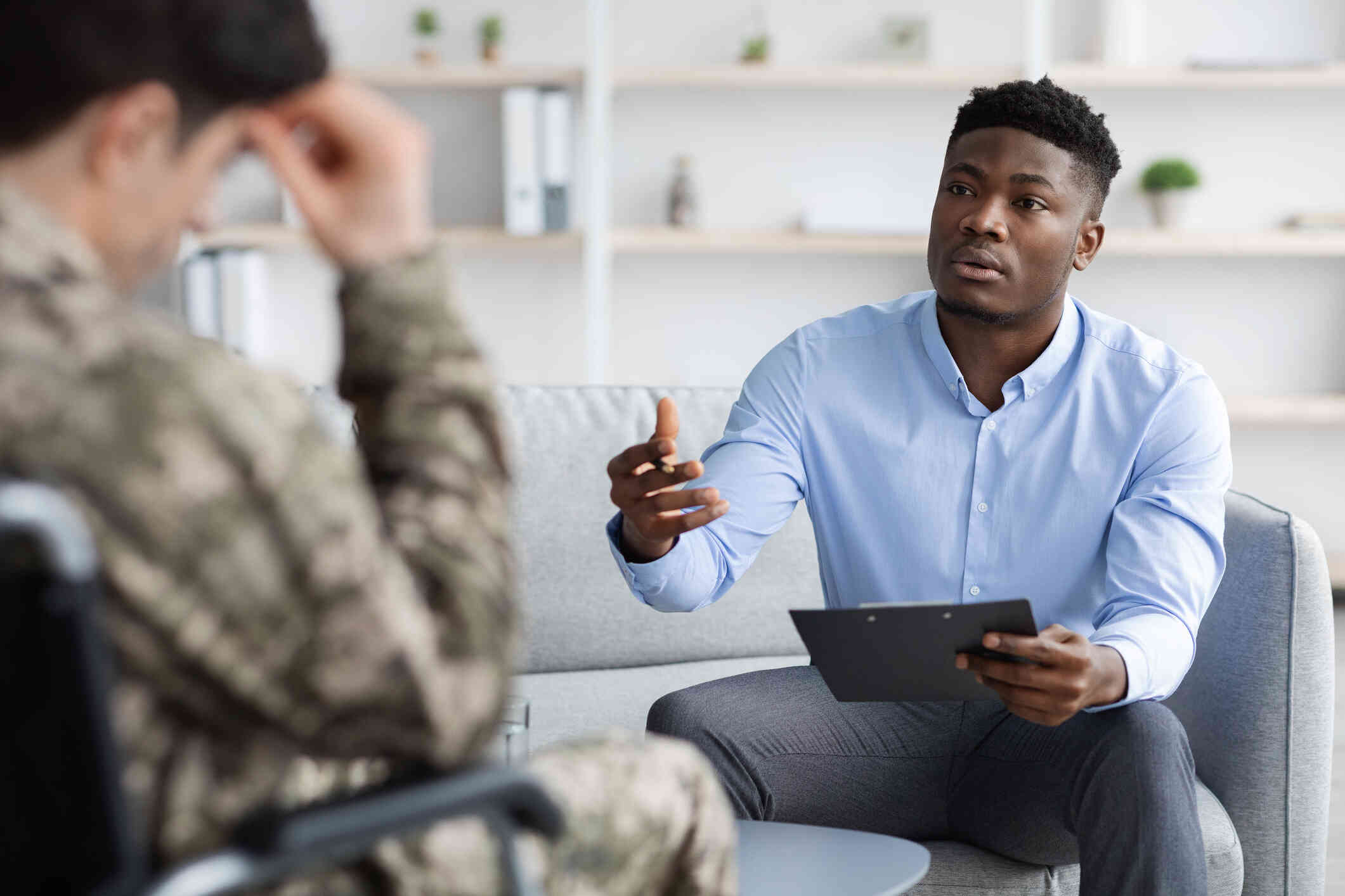 A male therapist holds a clipboard while talking to the patient in a military uniform sitting across from him during a therapy session.
